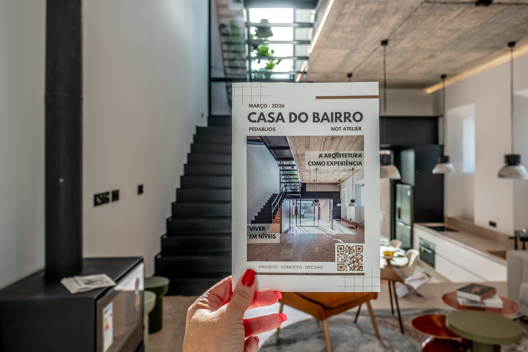 Débora segura a revista Casa do Bairro — Pedablios × Not Atelier — com o interior da casa ao fundo