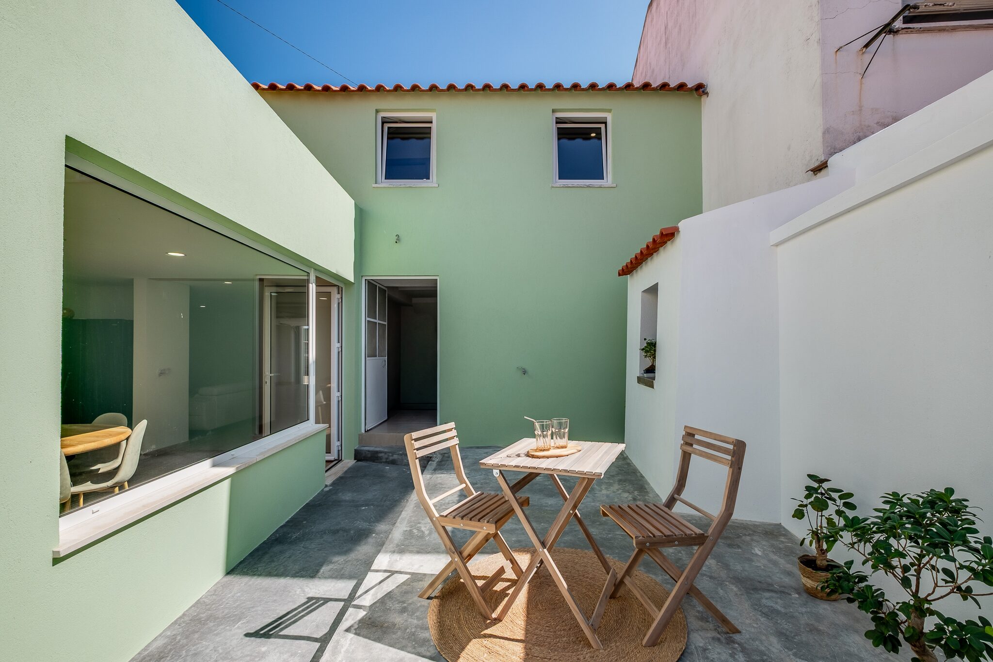 Pátio exterior com mesa e duas cadeiras ao sol, janelão da sala de jantar e muros em verde-menta