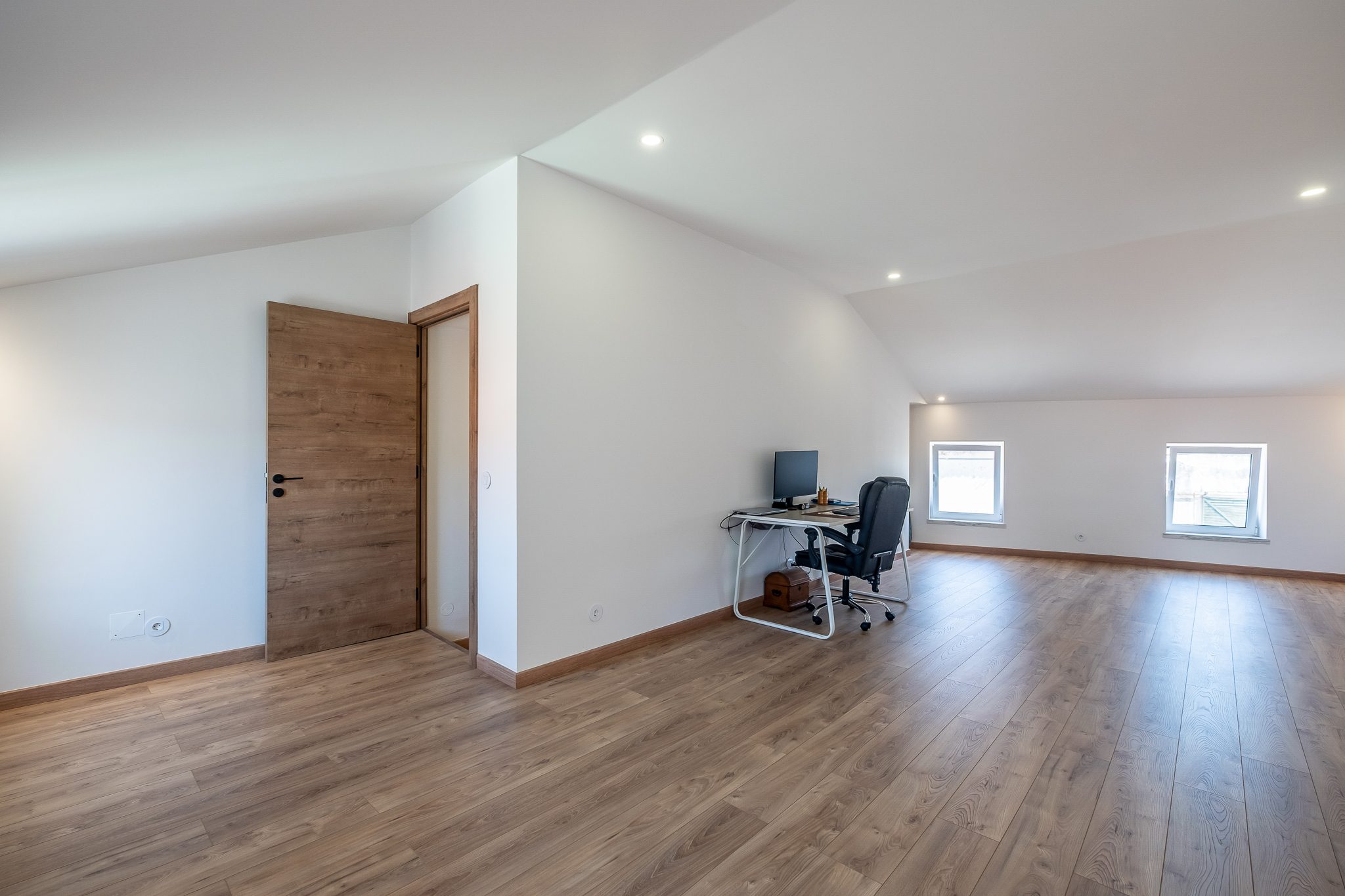 Quarto amplo sob teto inclinado, com soalho em madeira e zona de escritório